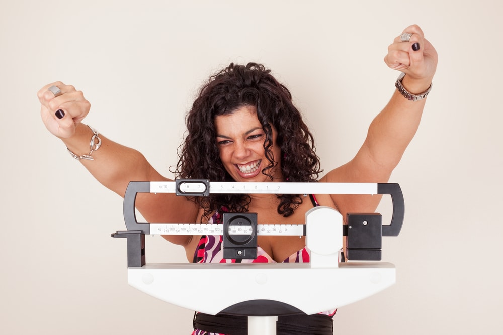 Woman on scale at doctor’s office with hands in the air, celebrating her weight loss.